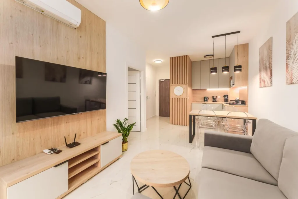 Salon z telewizorem na drewnianym panelu, szarą sofą i aneksem kuchennym w głębi apartamentu