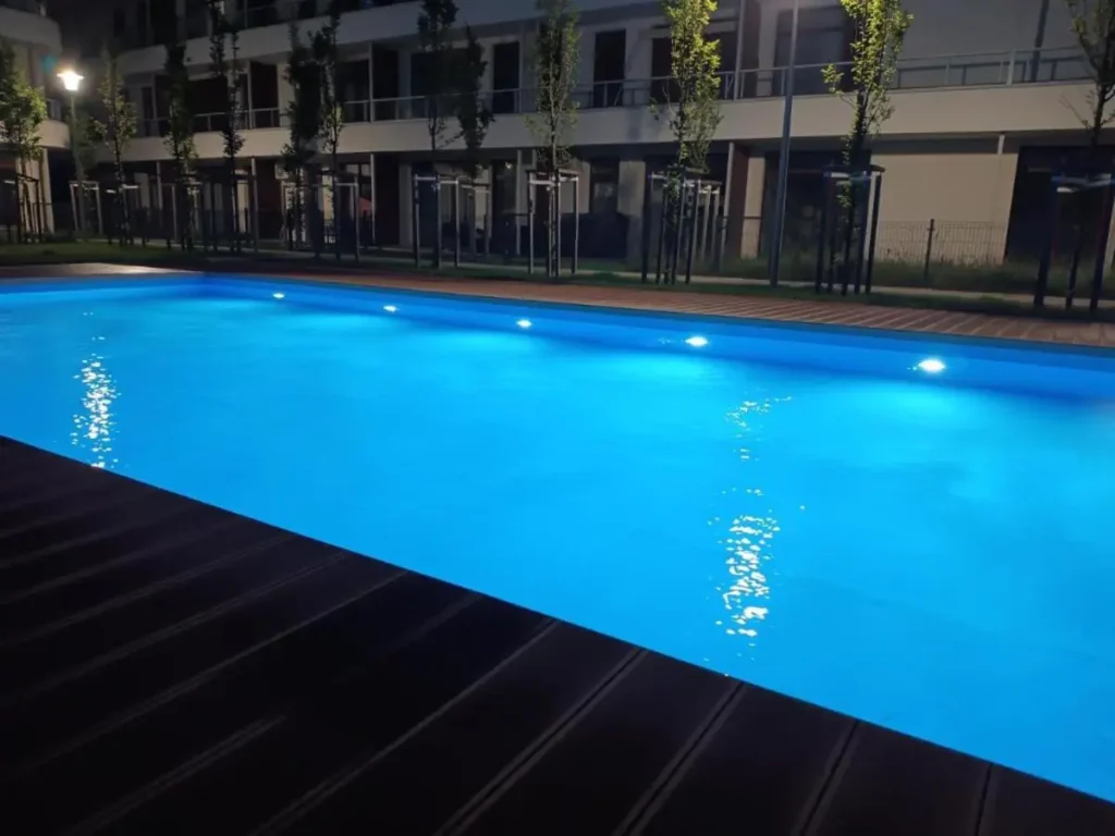 Podświetlony wieczorem basen odkryty na terenie osiedla, w tle budynek z apartamentami